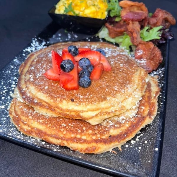 Stack of pancakes topped with strawberries and blueberries, served with bacon and scrambled eggs.