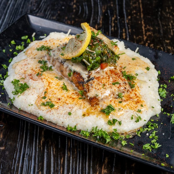 Grilled fish with creamy white sauce, greens, and lemon slices on a black plate.