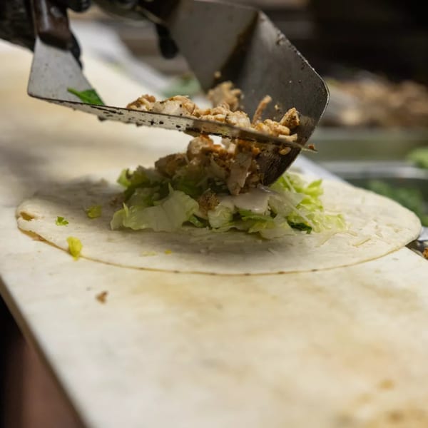A spatula adds seasoned chicken pieces to a tortilla with lettuce and sauce.