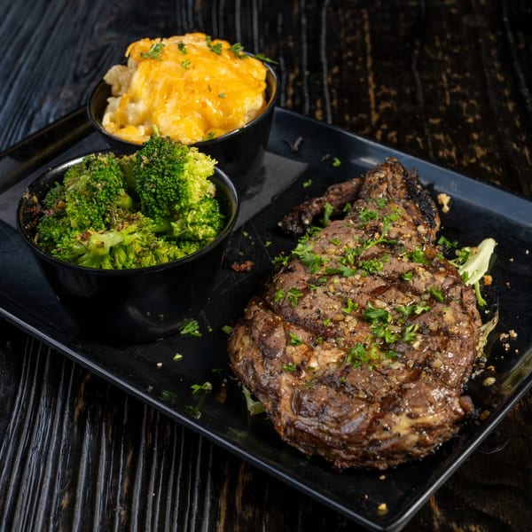 A grilled steak with parsley, two bowls of broccoli, and mac and cheese on a black tray.