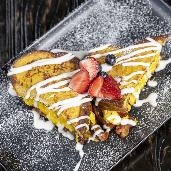 French toast with scrambled eggs, bacon, strawberries, blueberries, and powdered sugar on a black plate.