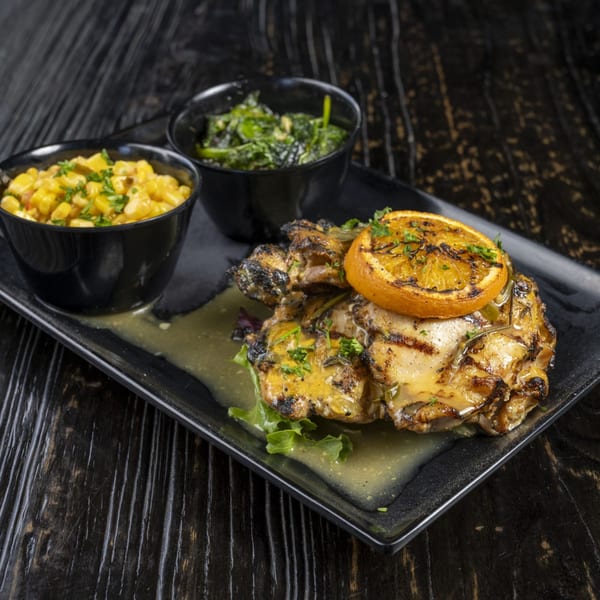 Grilled chicken with a charred orange slice, served with corn and greens in black bowls.