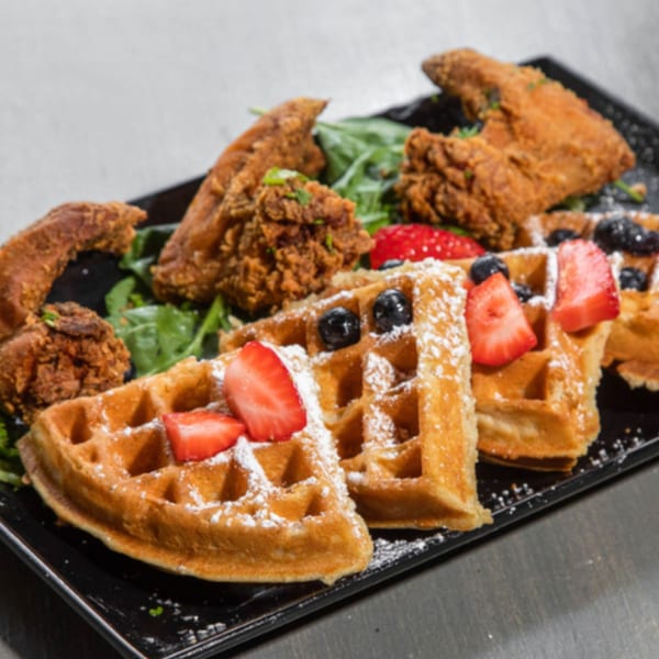 Crispy fried chicken and waffles with strawberries, blueberries, and powdered sugar on a black platter.