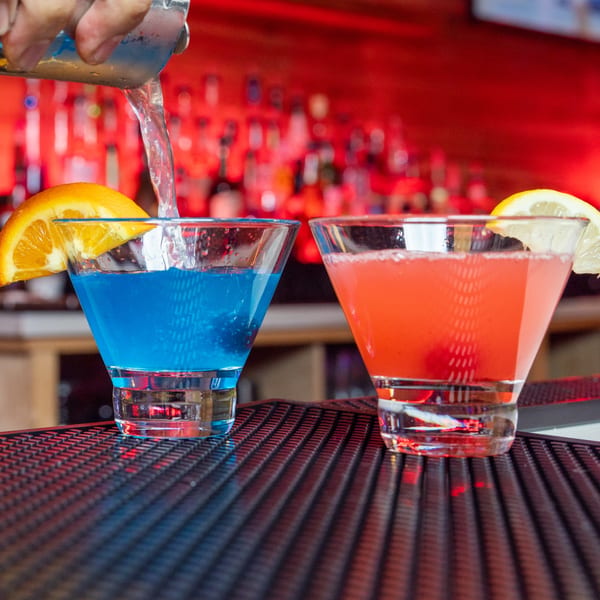 Bartender pouring a blue cocktail into a glass next to a red cocktail on a bar.